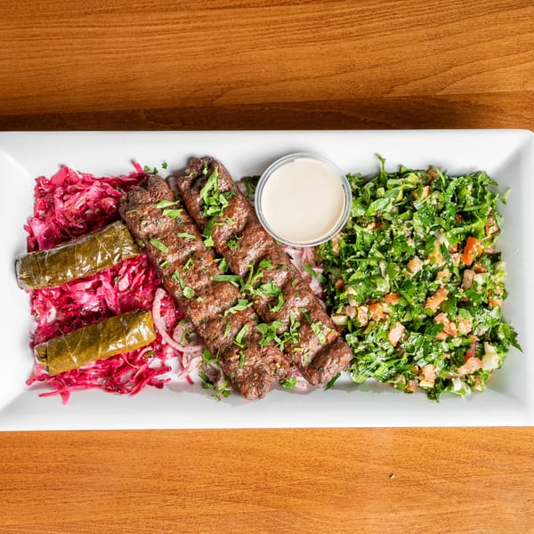 Two grilled meat skewers with pickled cabbage, stuffed grape leaves, tabbouleh salad, and tahini sauce.