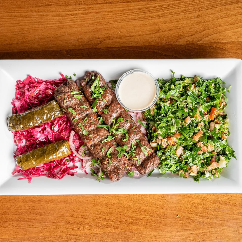 Two grilled meat skewers with pickled cabbage, stuffed grape leaves, tabbouleh salad, and tahini sauce.