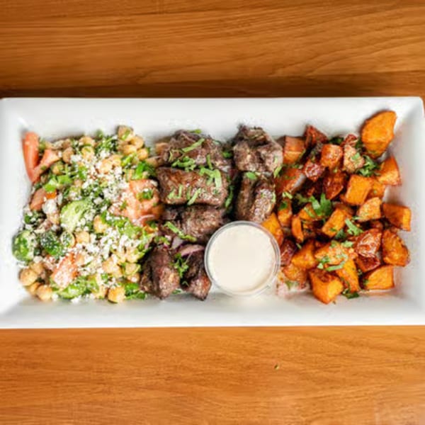 A white rectangular plate with steak bites, a chickpea salad, roasted sweet potatoes, and a dipping sauce.