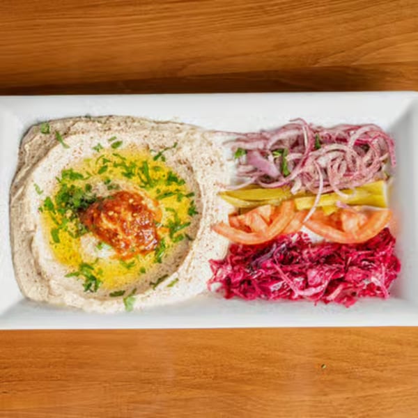 A white rectangular plate with hummus, olive oil, chili paste, red onions, pickles, tomatoes, and red cabbage.