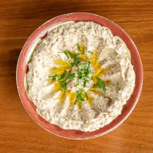 Creamy baba ghanoush topped with olive oil and fresh parsley in a red bowl on a wooden table.