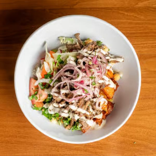 Overhead view of a white bowl filled with a colorful salad, meat, and dressing on a wooden table.