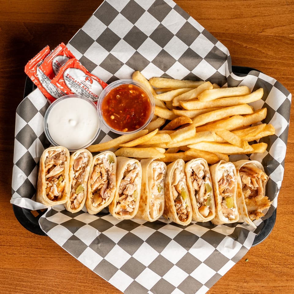 A basket of chicken shawarma wraps, golden fries, ketchup packets, and dipping sauces on checkered paper.
